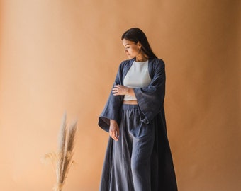May include: A woman wearing a matching set of blue-grey loungewear. The set includes a long, open-front robe with wide sleeves, a cropped top, and wide-leg pants. A vase with tall grass and a matching belt are on the floor.