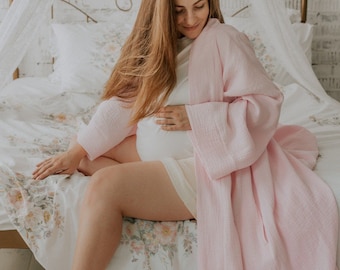 May include: A pregnant woman wearing a pink robe sits on a bed with a floral patterned bedspread. She is holding her belly with one hand and her other hand is resting on the bed.