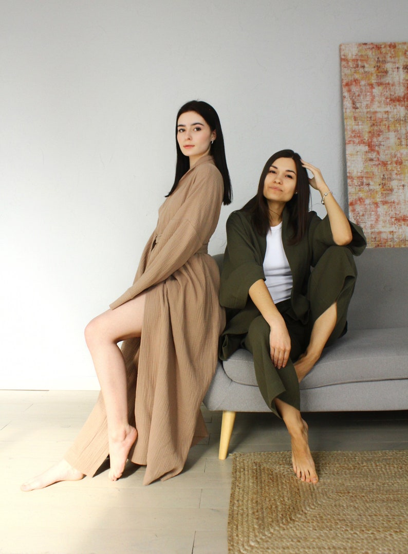 May include: Two women wearing long robes, one in a light brown robe and the other in a dark green robe. The woman in the brown robe is standing and the woman in the green robe is sitting on a grey sofa.
