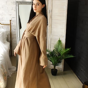 Pode incluir: Uma mulher vestindo um longo robe marrom com textura de waffle e uma faixa amarrada na cintura. Ela está em pé em um quarto com paredes de tijolo branco e uma tela de madeira branca atrás dela.