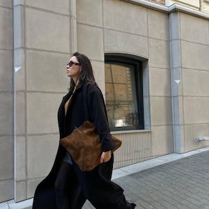 Może przedstawiać: Kobieta w czarnym płaszczu i czarnych spodniach idzie chodnikiem. Niesie brązową skórzaną torbę.