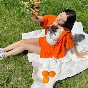May include: A person wearing an orange shirt and shorts sits on a white blanket in a grassy area. They are holding a bunch of carrots and there are three oranges on the blanket.
