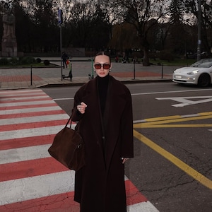 Puede incluir: Una mujer con un abrigo marrón, un jersey de cuello alto negro y gafas de sol se encuentra en un paso de cebra. Lleva un bolso de mano de cuero marrón.