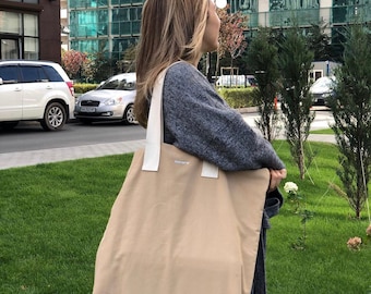 May include: A beige tote bag with white straps. The bag has a small logo on the front that says "Something".