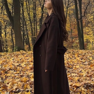 Könnte beinhalten: Ein langer, dunkelbrauner Mantel mit einem Gürtel in der Taille. Der Mantel wird in einer Waldlandschaft mit gelben Herbstblättern getragen. Die Person blickt nach oben und schließt die Augen.