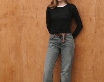 May include: A woman wearing a black long-sleeved shirt, blue jeans, and brown loafers. She is standing in front of a wood panel wall.