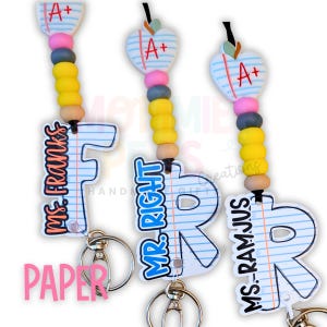 May include: Three keychains with a school theme. Each keychain features a letter-shaped tag with a lined paper design and a name. The keychains have a pencil-themed bead design and an apple-shaped charm with an "A+" grade.