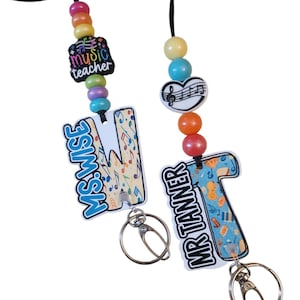May include: Two personalized music teacher lanyards. One features the text "MS. WISE" on a letter "M" with musical notes, and the other says "MR TANNER" on a letter "T" with musical instruments. Both have colorful beads and key rings.
