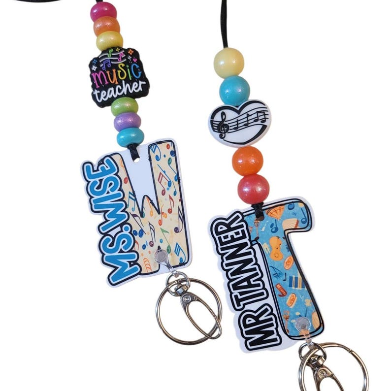 May include: Two personalized music teacher lanyards. One features the text "MS. WISE" on a letter "M" with musical notes, and the other says "MR TANNER" on a letter "T" with musical instruments. Both have colorful beads and key rings.