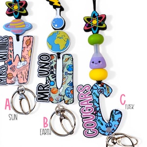 May include: Three science-themed lanyards with keychains. One lanyard features a planet design, another has a globe, and the third has a flask. Each lanyard has a letter and a science-related charm. The lanyards are in various colors.