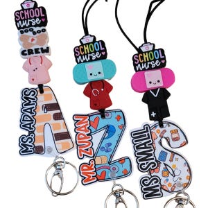May include: Three personalized badge reels for school nurses. Each reel features a name, a decorative band-aid, and a miniature nurse's uniform. The reels are attached to a black lanyard with a metal clip.