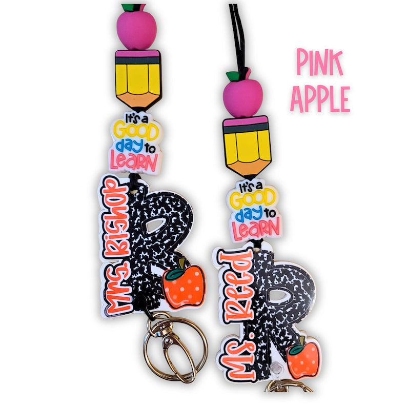 May include: Two teacher lanyards with a pink apple, yellow pencil, and the text "It's a good day to learn." One lanyard says "Mrs. Bishop" and the other says "Ms. Reed." Each has a black and white patterned letter "R" and a small orange apple.