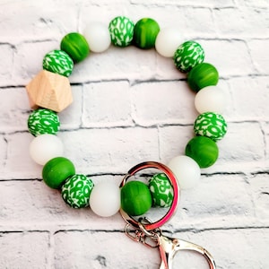 May include: A green and white beaded keychain wristlet. The wristlet features a mix of solid green, white, and patterned green beads, along with a wooden geometric bead. A silver-colored clasp and key ring are attached.