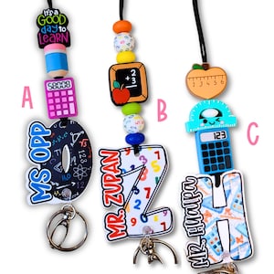 May include: Three teacher-themed lanyards with keychains. Each lanyard features a different design: a pencil and calculator, a chalkboard and apple, and a ruler and calculator. The lanyards have colorful beads and the text "It's a good day to learn".