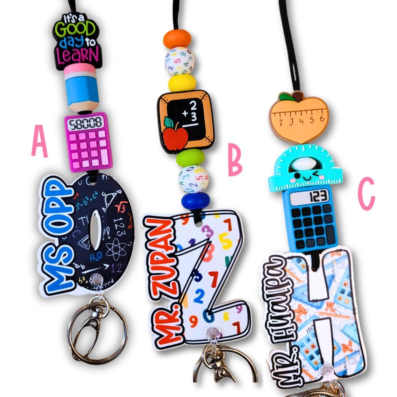 May include: Three teacher-themed lanyards with keychains. Each lanyard features a different design: a pencil and calculator, a chalkboard and apple, and a ruler and calculator. The lanyards have colorful beads and the text "It's a good day to learn".