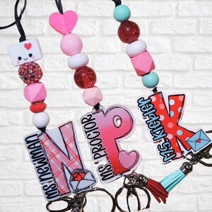 May include: Three beaded lanyards with personalized name tags. The lanyards feature pink, red, and white beads, with a variety of shapes. Each lanyard has a name tag with a teacher's name and a small envelope graphic. One lanyard has a blue tassel.