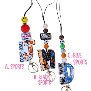 May include: Three sports-themed lanyards with keychains. One lanyard has a red apple, a smiley face bead, and the text "PHYSICAL EDUCATION teacher". Another has the text "COACH love-inspire". The third has the text "MR. DIME".