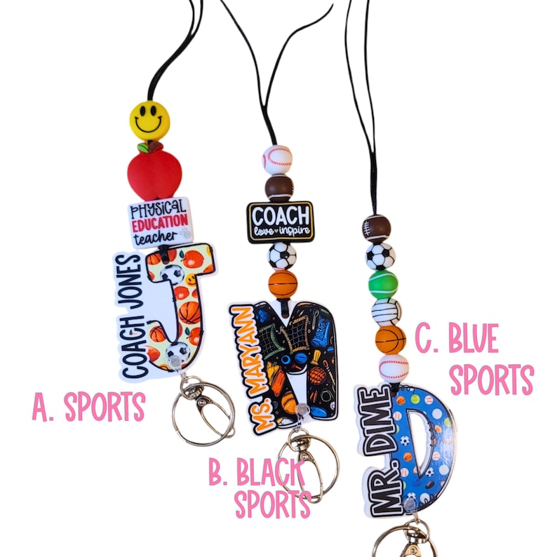 May include: Three sports-themed lanyards with keychains. One lanyard has a red apple, a smiley face bead, and the text "PHYSICAL EDUCATION teacher". Another has the text "COACH love-inspire". The third has the text "MR. DIME".