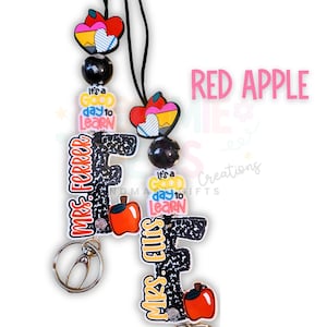 May include: A lanyard with a black cord, featuring colorful charms. The charms include a red apple, a heart with a pencil design, and the text "It's a good day to learn." The lanyard also has the text "MRS. FERRER" and "MRS. ELLIS".