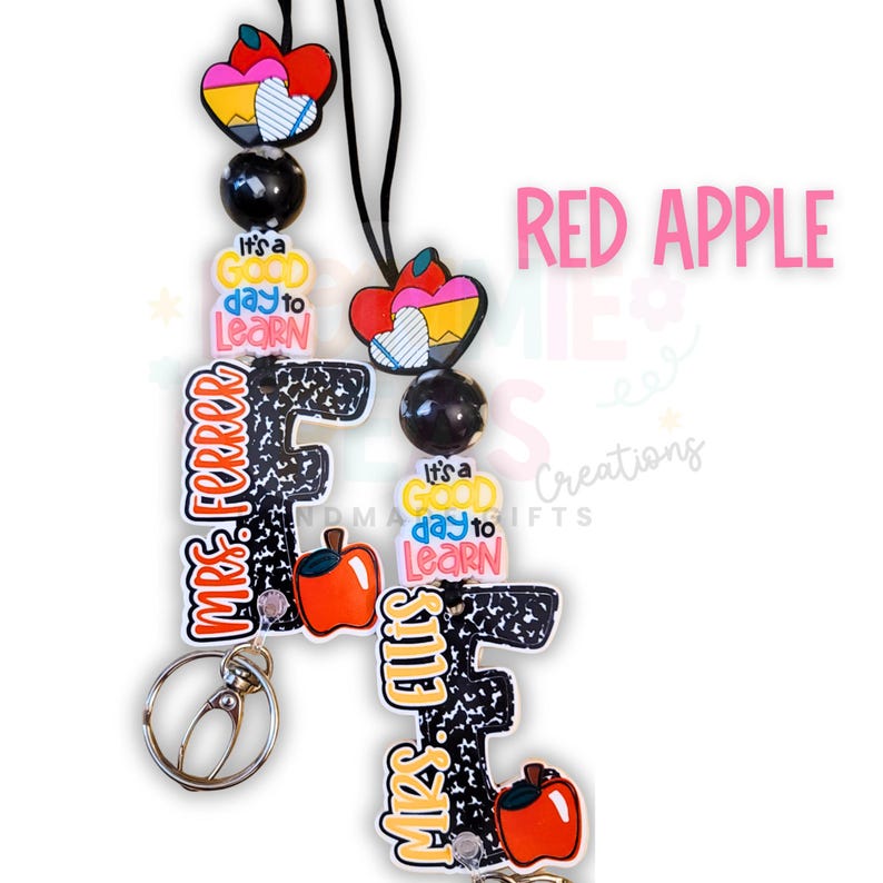 May include: A lanyard with a black cord, featuring colorful charms. The charms include a red apple, a heart with a pencil design, and the text "It's a good day to learn." The lanyard also has the text "MRS. FERRER" and "MRS. ELLIS".