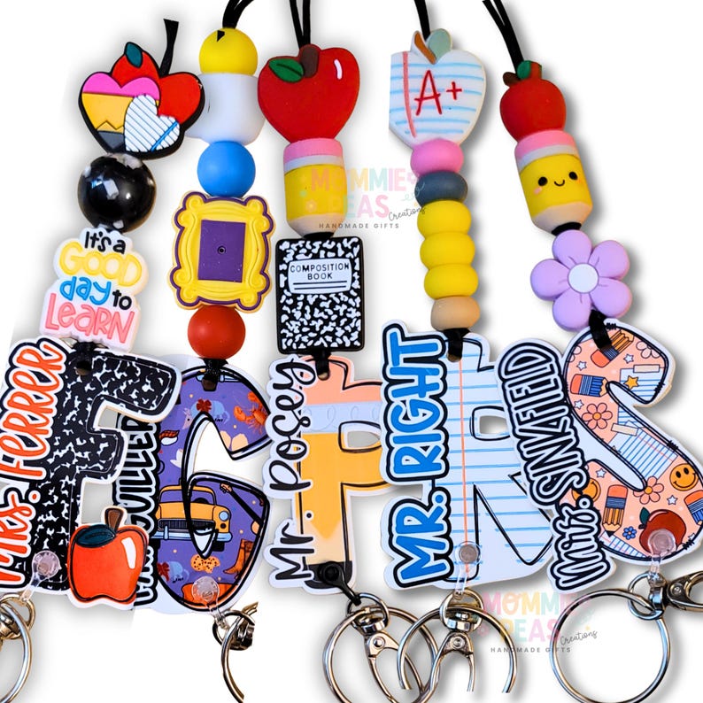 A collection of personalized teacher lanyards from Mommie Peas Creations featuring custom acrylic name charms, colorful silicone beads, and school-themed icons like pencils, composition books, and paper. Each lanyard includes a breakaway safety clasp and lobster clip for ID badges.