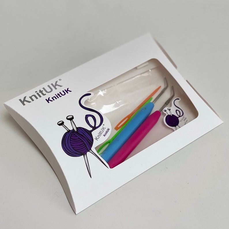 K&ouml;nnte beinhalten: Eine wei&szlig;e Verpackung mit einem durchsichtigen Fenster, das einen Satz bunter H&auml;kelnadeln zeigt. Die Verpackung ist mit "KnitUK" beschriftet und zeigt eine violette Wollkugel mit zwei Stricknadeln.