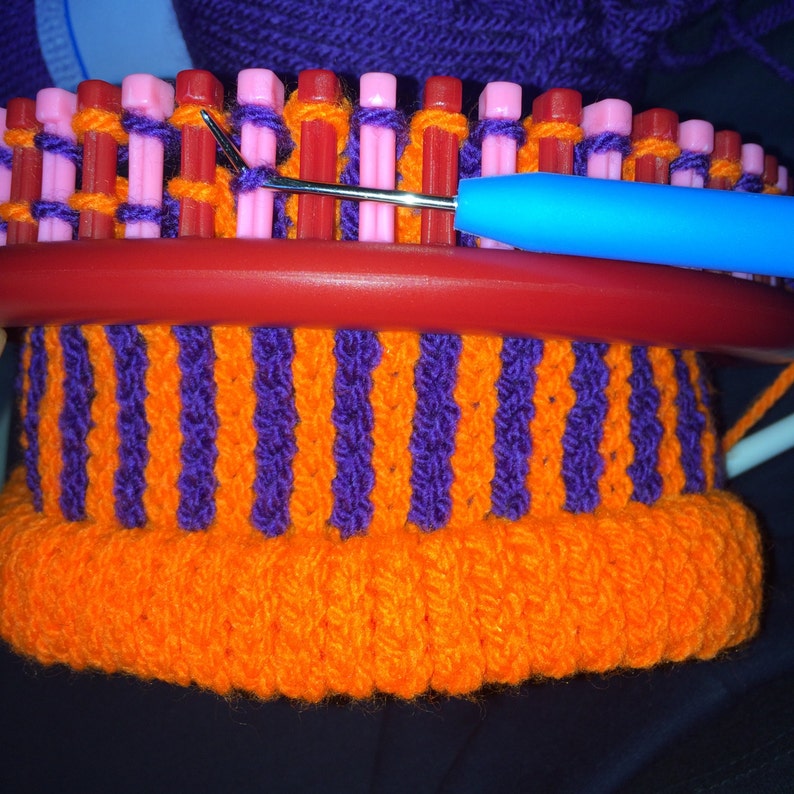 Puede incluir: Primer plano de un telar de punto con hilo naranja y morado. El hilo se est&aacute; tejiendo en un patr&oacute;n a rayas. Se est&aacute; utilizando una aguja de tejer para trabajar el hilo.