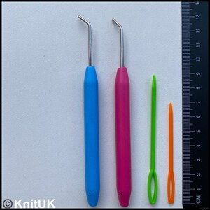 K&ouml;nnte beinhalten: Vier H&auml;kelnadeln, zwei mit blauen und rosa Griffen und zwei mit gr&uuml;nen und orangefarbenen Griffen. Die H&auml;kelnadeln sind neben einem Lineal ausgerichtet, das 14 cm misst.