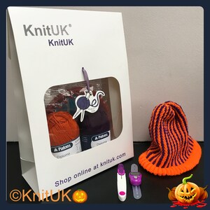May include: A white box with a clear window showing orange and purple yarn, knitting needles, and a tag that says "KnitUK". The box also has the text "Shop online at knituk.com". There is a purple and orange striped knitted hat, two purple and white knitting needles, and a purple and white stitch counter on a white surface.