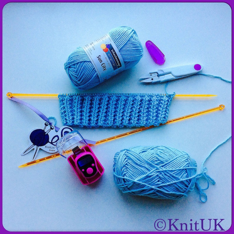 May include: A blue knitting project with two balls of yarn, knitting needles, a stitch counter, and a pair of scissors. The yarn is labeled "Schachenmayr - Original Sun City".