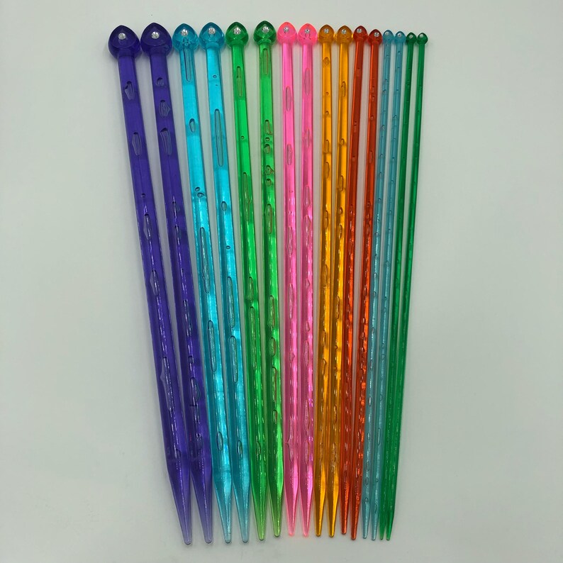 May include: A set of 14 colorful plastic knitting needles with heart-shaped tops. The needles are arranged in a row from shortest to longest. The colors include purple, blue, green, pink, orange, and yellow.