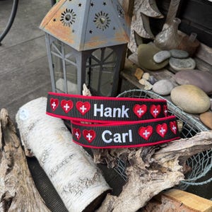 Könnte beinhalten: Zwei schwarze Hundehalsbänder mit rotem Rand, bestickt mit den Namen "Hank" und "Carl" in Weiß. Jedes Halsband zeigt ein sich wiederholendes Muster aus roten Herzen und weißen Kreuzen, ein Schweizer Design.