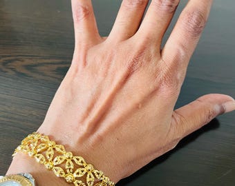 May include: A gold-colored bracelet with an openwork design is worn on a wrist. The bracelet features a repeating pattern of floral or leaf-like shapes. A gold and silver watch is also visible.