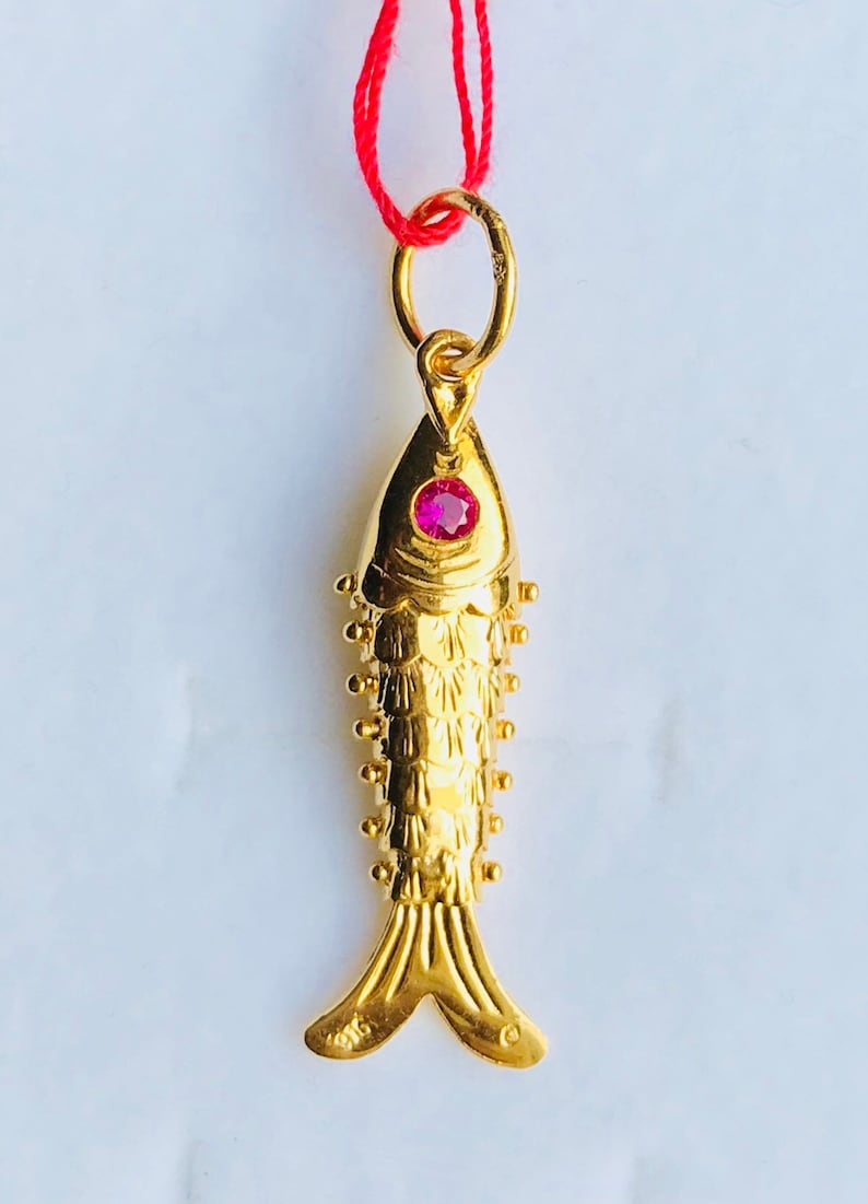 May include: A gold fish pendant with a red gemstone eye. The pendant is on a red string.