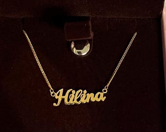 May include: A gold-colored necklace featuring a nameplate pendant with the name Hilina written in a cursive script font. The pendant is attached to a delicate gold-colored chain that extends outward from both sides of the nameplate.
