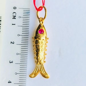 May include: A gold fish pendant with a red gemstone eye. The pendant is approximately 3 cm long and is hanging from a red string.