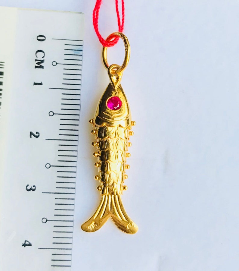 May include: A gold fish pendant with a red gemstone eye. The pendant is approximately 3 cm long and is hanging from a red string.