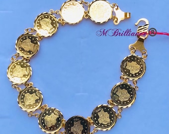 Puede incluir: Una pulsera de tono dorado compuesta por once dijes circulares en forma de moneda, cada uno con un retrato de perfil y el año 1906. Los eslabones están conectados por pequeños anillos dorados y la pieza incluye un cierre de gancho con el texto MBrilliance.