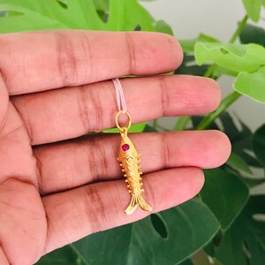 May include: A gold-colored fish pendant with a red gemstone eye. The pendant is hanging from a white string.