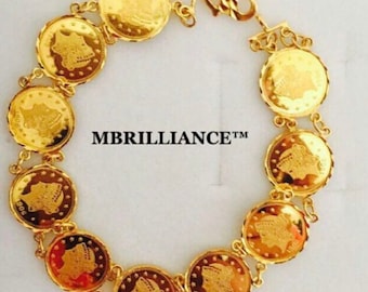 Puede incluir: Una pulsera de tono dorado compuesta por once dijes circulares en forma de moneda unidos por pequeños anillos dorados. Cada dije presenta un retrato de perfil y el texto MBRILLIANCE™ se encuentra centrado dentro de la disposición circular.