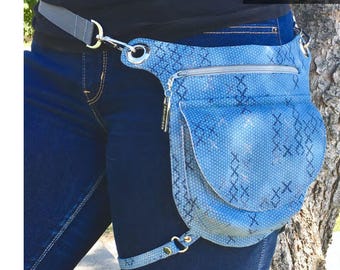 May include: A light blue fabric holster bag with a zipped pocket and a flap pocket. The bag is worn on a person's hip and secured with a belt. The bag is 19 cm wide and 23 cm tall. The text "Charlie Holster Bag" is at the top of the image. The text "Design by Tara Sinclair" is at the bottom of the image. The text "Uh Oh Creation" and "Serial Bagmakers" is in the bottom right corner of the image.