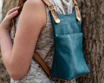May include: A teal blue leather backpack with brown straps and gold hardware. The backpack has a flap closure and a small pocket on the front. The backpack is being worn by a person.