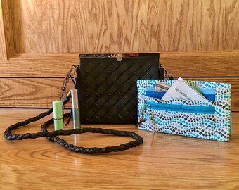 May include: A black woven shoulder bag with a silver clasp and a black braided strap. The bag is sitting on a wooden surface with a small blue and white patterned wallet and two tubes of cosmetics.