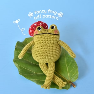 May include: A green crocheted frog wearing a red and white crocheted mushroom hat sits on a green leaf. The frog has black eyes and a white smile. The background is a light blue.