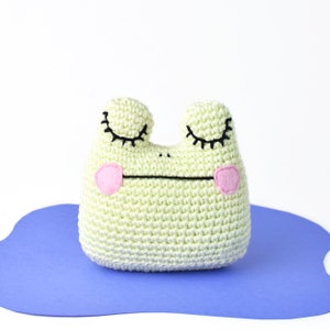 May include: A light green crocheted frog with black eyes and pink cheeks. The frog is lying on a blue surface.