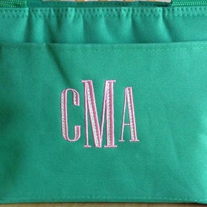 K&ouml;nnte beinhalten: Eine gr&uuml;ne Einkaufstasche mit einem gestickten Monogramm in Rosa und Wei&szlig;, das "CMA" lautet.
