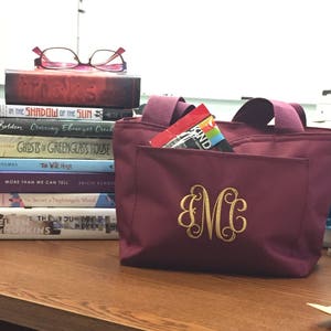 K&ouml;nnte beinhalten: Eine bordeauxfarbene Tragetasche mit einem goldenen Monogramm "MC", das auf der Vorderseite gestickt ist. Die Tasche hat eine Rei&szlig;verschlusstasche und steht auf einer Holzoberfl&auml;che mit einem Stapel B&uuml;cher dahinter.