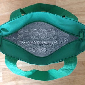 K&ouml;nnte beinhalten: Eine gr&uuml;ne, isolierte Lunch-Tasche mit Rei&szlig;verschluss und zwei Griffen. Die Tasche ist offen und das Innere ist mit einem silbernen reflektierenden Material ausgekleidet.