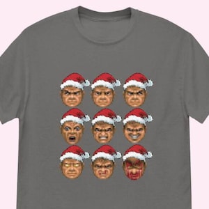 May include: A gray t-shirt featuring a grid of nine pixelated faces, each wearing a Santa hat. The faces display various expressions, ranging from angry to surprised. The design is a humorous take on a classic video game.