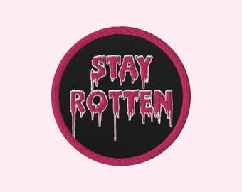 Może przedstawiać: Haftowana naszywka z napisem "STAY ROTTEN" w różowych, ociekających literach na czarnym, okrągłym tle. Naszywka ma różową obwódkę i jest umieszczona na jasnoróżowym tle. Idealna do personalizacji odzieży lub akcesoriów.